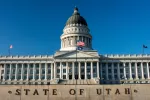 The front of the Utah capitol with a bright blue sky with the words State of Utah in the foreground. 