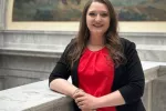Dark haired woman wearing a red blouse leans on a granite railing at the capitol.