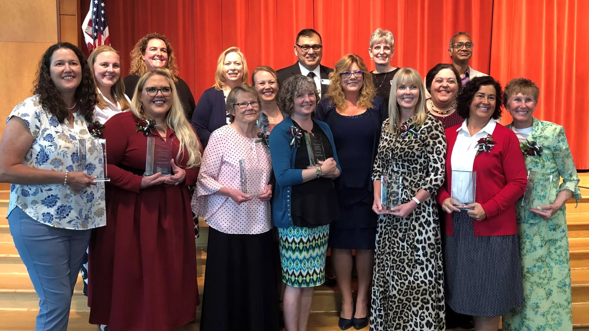 15 people stand together in dresses and suits holding trophies and smiling.