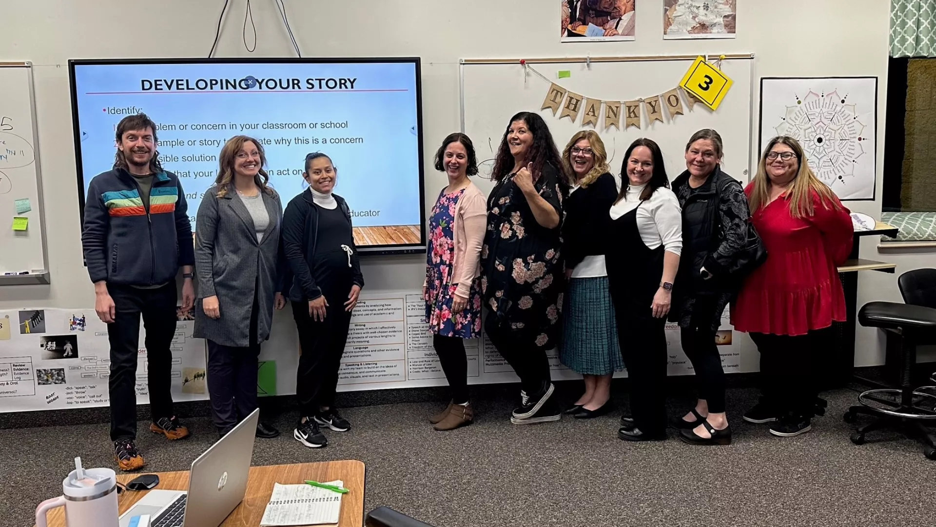 Nine people standing against a classroom wall smiling at the camera.