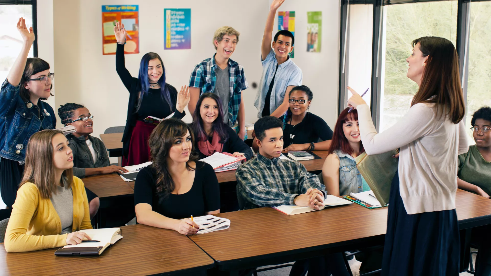 Female teacher in cardigan interacts with group of high school students of various races and genders.