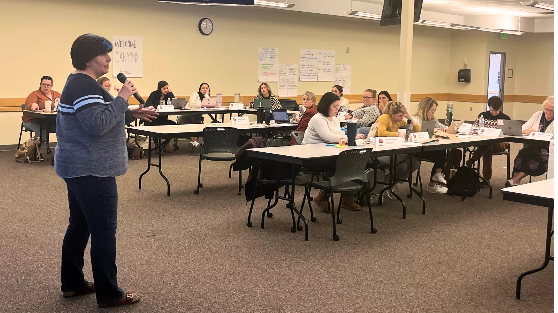 Woman wearing sweater in jeans holds microphone while speaking to group of educators sitting in rows of tables. 