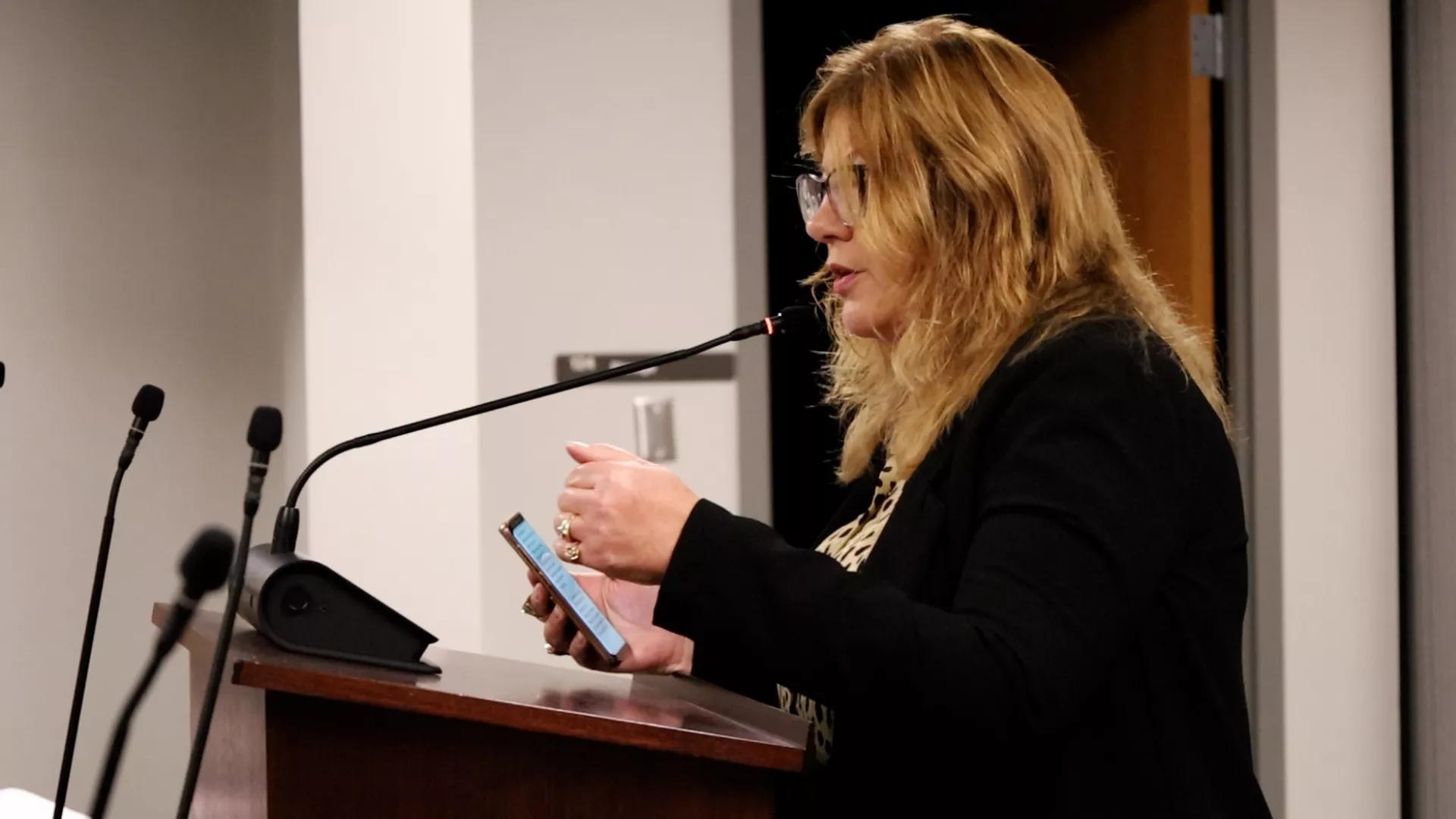 President Renee Pinkney stands at podium speaking to Utah State Board of Education with phone in her hand. 