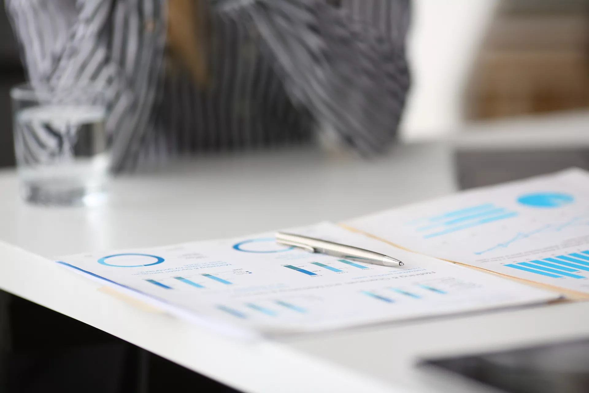 Papers on a table with a person's elbows and a glass of water in the background. 