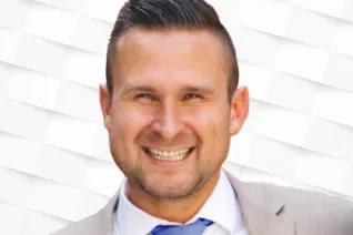 White man against a white background smiling wearing a suit and tie.