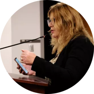 President Renee Pinkney stands at podium speaking to Utah State Board of Education with phone in her hand.