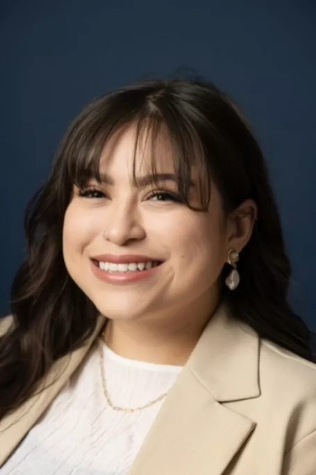 Woman smiles with a dark blue background and a tan jacket.