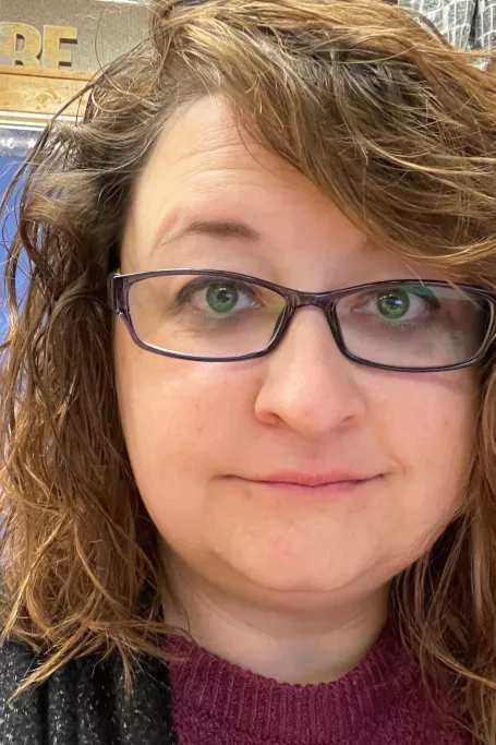 Woman with curly hair and glasses looks at the camera in a classroom.