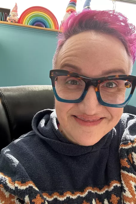 White woman in a blue sweater smiling at the camera. She has pink hair and trendy glasses.