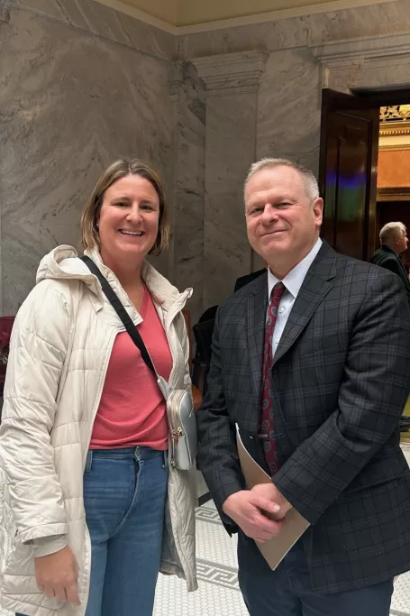 White woman with blonde hair and cream coat smiles next to lawmaker at the capitol. 