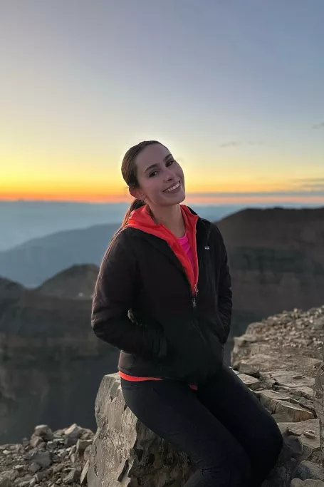 Woman sitting near a cliff with a sunset over a rock vista.