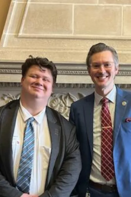 Two men in suits stand in front of stone fireplace.
