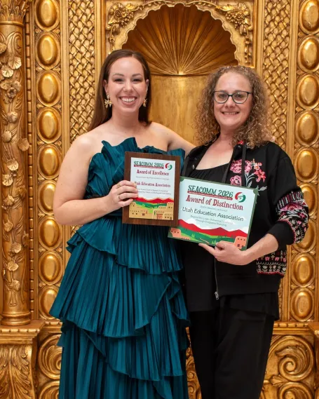 Woman in teal gown and woman in black hoodie smile holding awards in front of gold background.