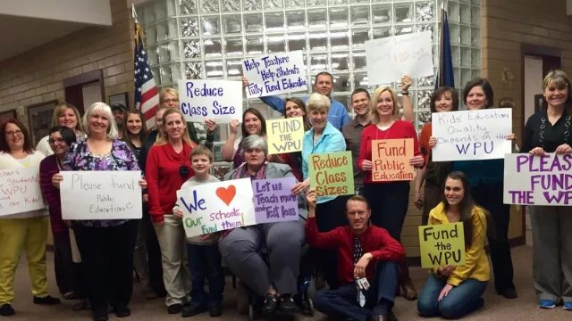 Educators hold signs advocating for school funding in a school atrium.
