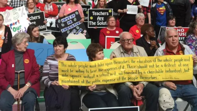 Retired educators sit with a sign in a large crowd rallying for public education at the state capitol.