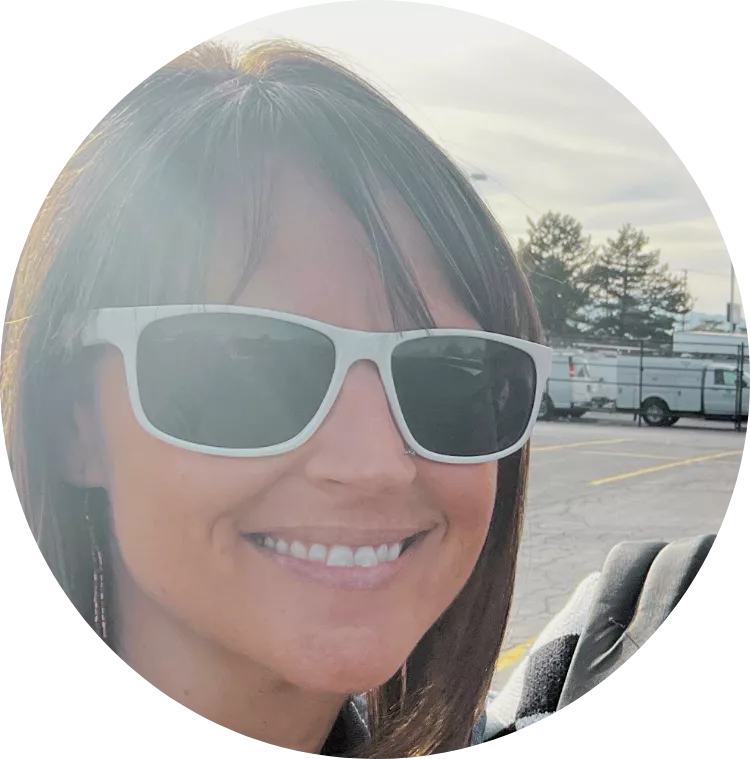 White woman with brown hair smiling with white-rimmed sunglasses outside in a parking lot